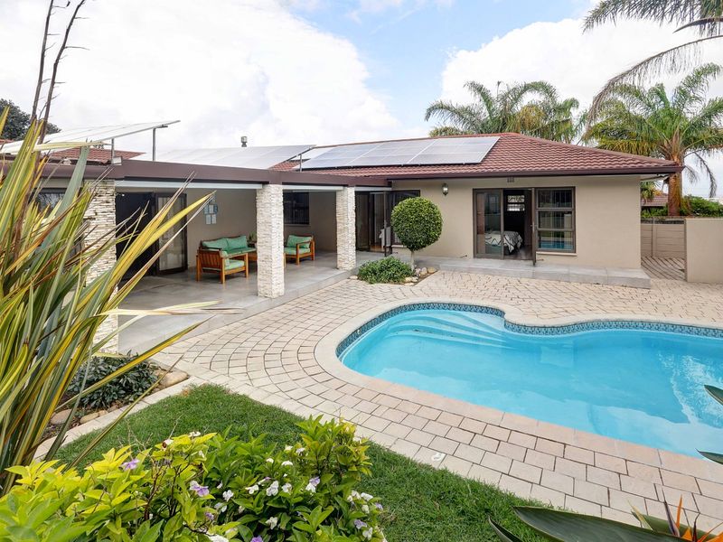 A well-maintained outdoor entertainment area featuring a swimming pool, paved deck, covered patio with stone pillars, and roof-mounted solar panels.