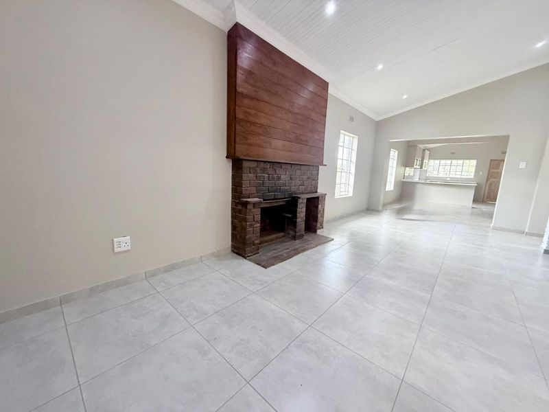 Spacious open-plan living area featuring a prominent brick fireplace with a wooden mantle, neutral tiled flooring, and a vaulted ceiling leading to the kitchen.