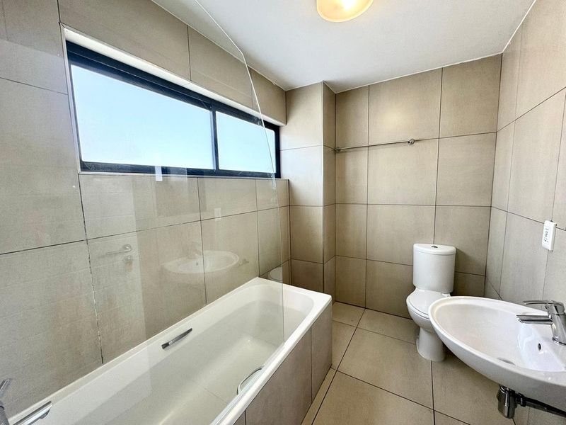 A clean, modern bathroom featuring neutral floor-to-ceiling tiling, a bathtub with a glass screen, a pedestal basin, and a toilet.