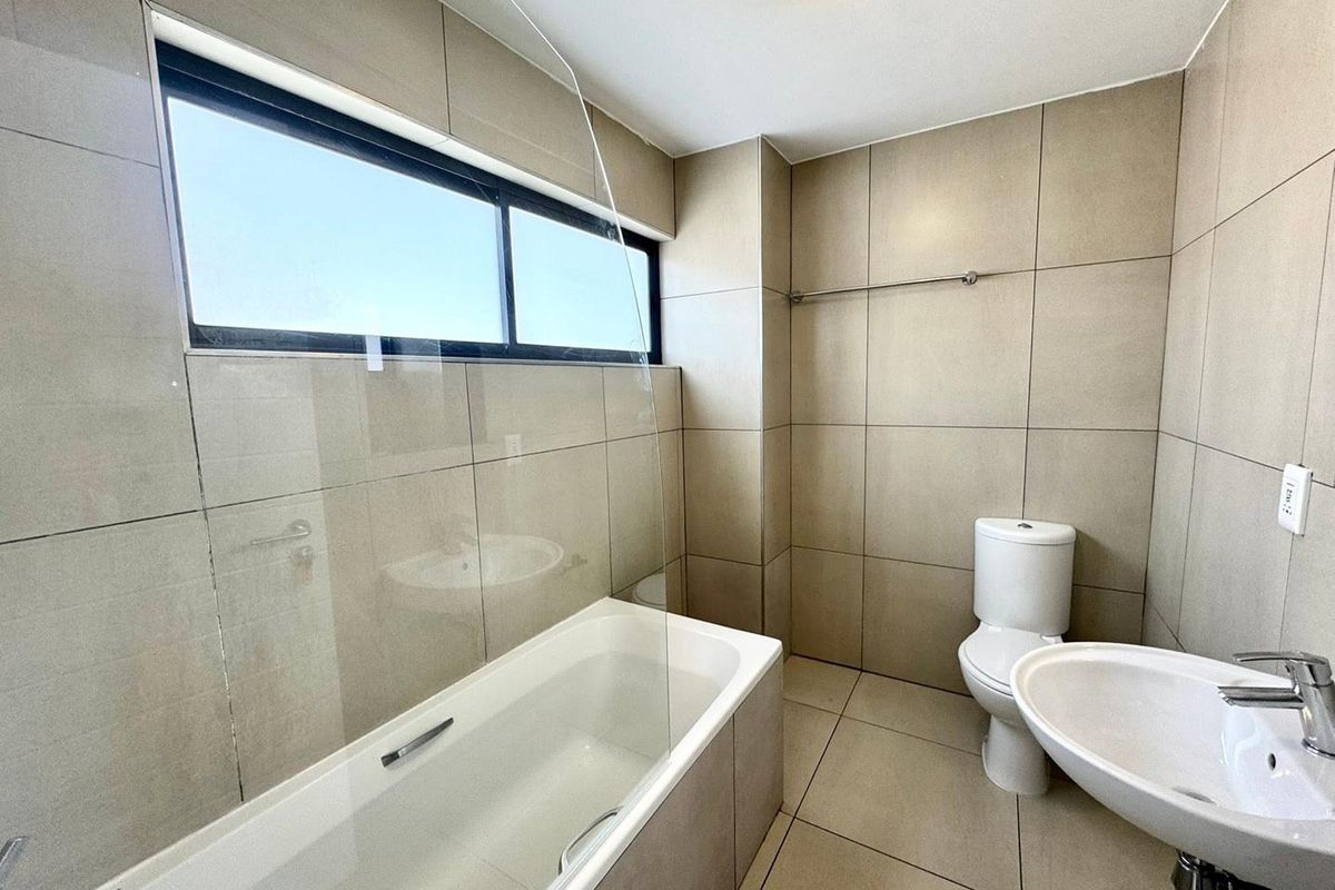 A clean, modern bathroom featuring neutral floor-to-ceiling tiling, a bathtub with a glass screen, a pedestal basin, and a toilet.