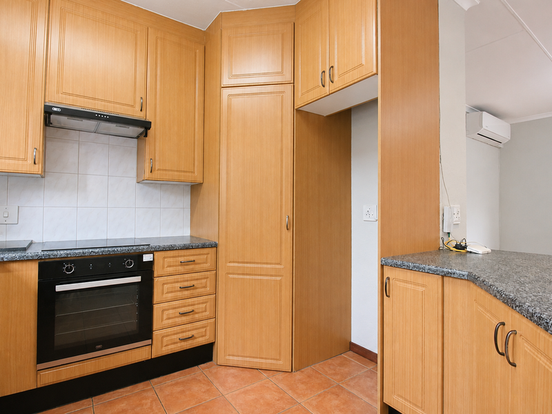 A functional kitchen featuring light wood-finish cabinetry, granite-look countertops, built-in oven, electric hob, and tiled flooring, with an adjacent air-conditioned living space.