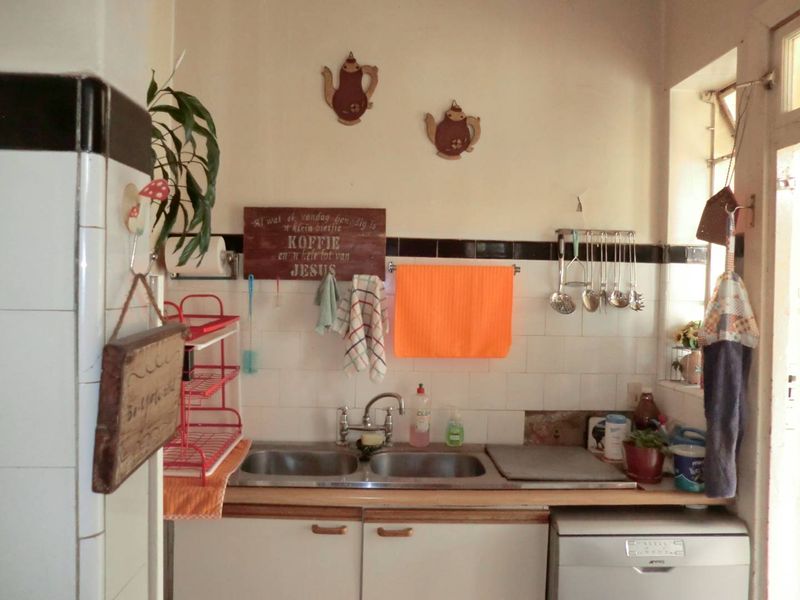 A compact kitchen area featuring a double bowl stainless steel sink, tiled splashback, and base cabinetry, showing signs of wear and missing tiles.
