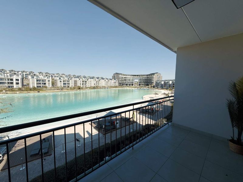 A spacious balcony featuring tiled flooring and a metal balustrade, offering expansive views over the estate's lagoon and surrounding apartment buildings.