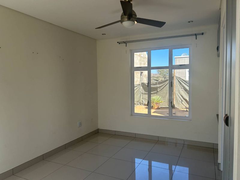 A bright, empty bedroom featuring neutral tiled flooring, a ceiling fan, and a large window with a curtain rod, ready for personalization.