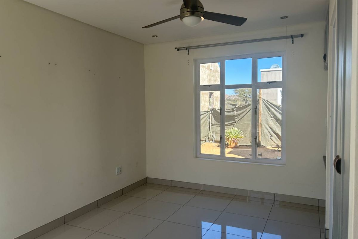 A bright, empty bedroom featuring neutral tiled flooring, a ceiling fan, and a large window with a curtain rod, ready for personalization.