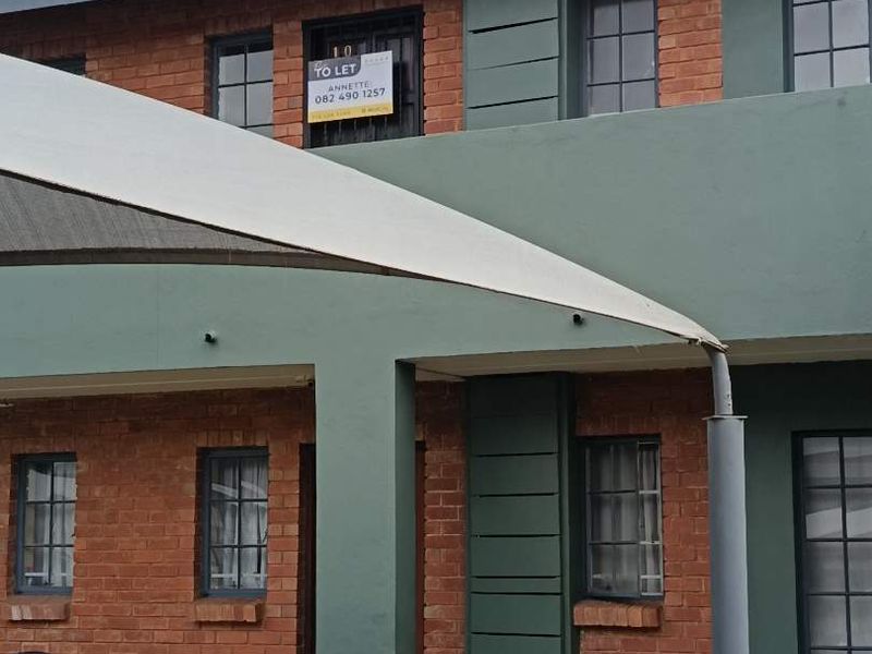 A two-story apartment building exterior featuring a brick facade, tiled roof, and a ground-floor patio area with a shade sail.