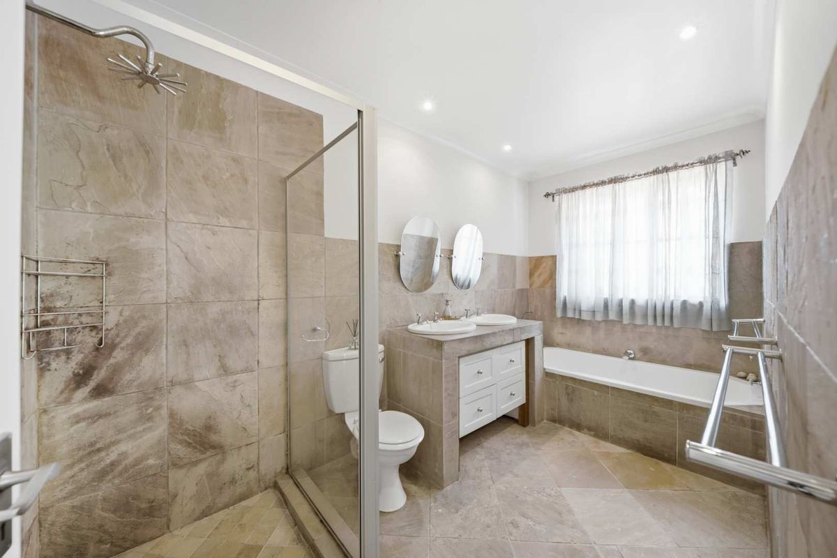 A well-appointed bathroom featuring a double vanity, built-in bathtub, separate walk-in shower, and neutral stone-look tiling throughout.