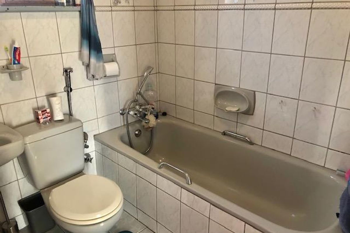 A dated bathroom featuring a standard bathtub with shower mixer, wall-mounted toilet, and full-height ceramic wall tiling.