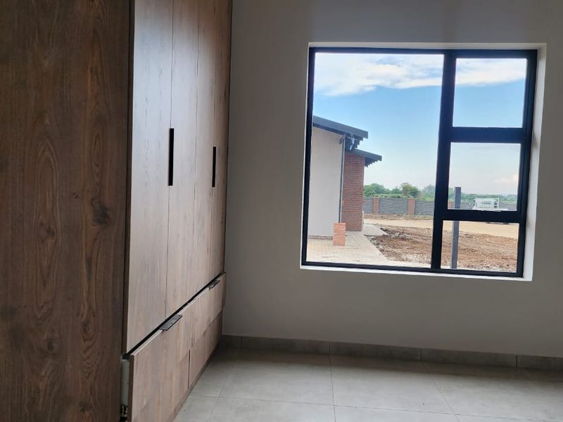 A bright, modern bedroom featuring neutral tiled floors, built-in wooden cupboards, and a large window with views of the exterior grounds.