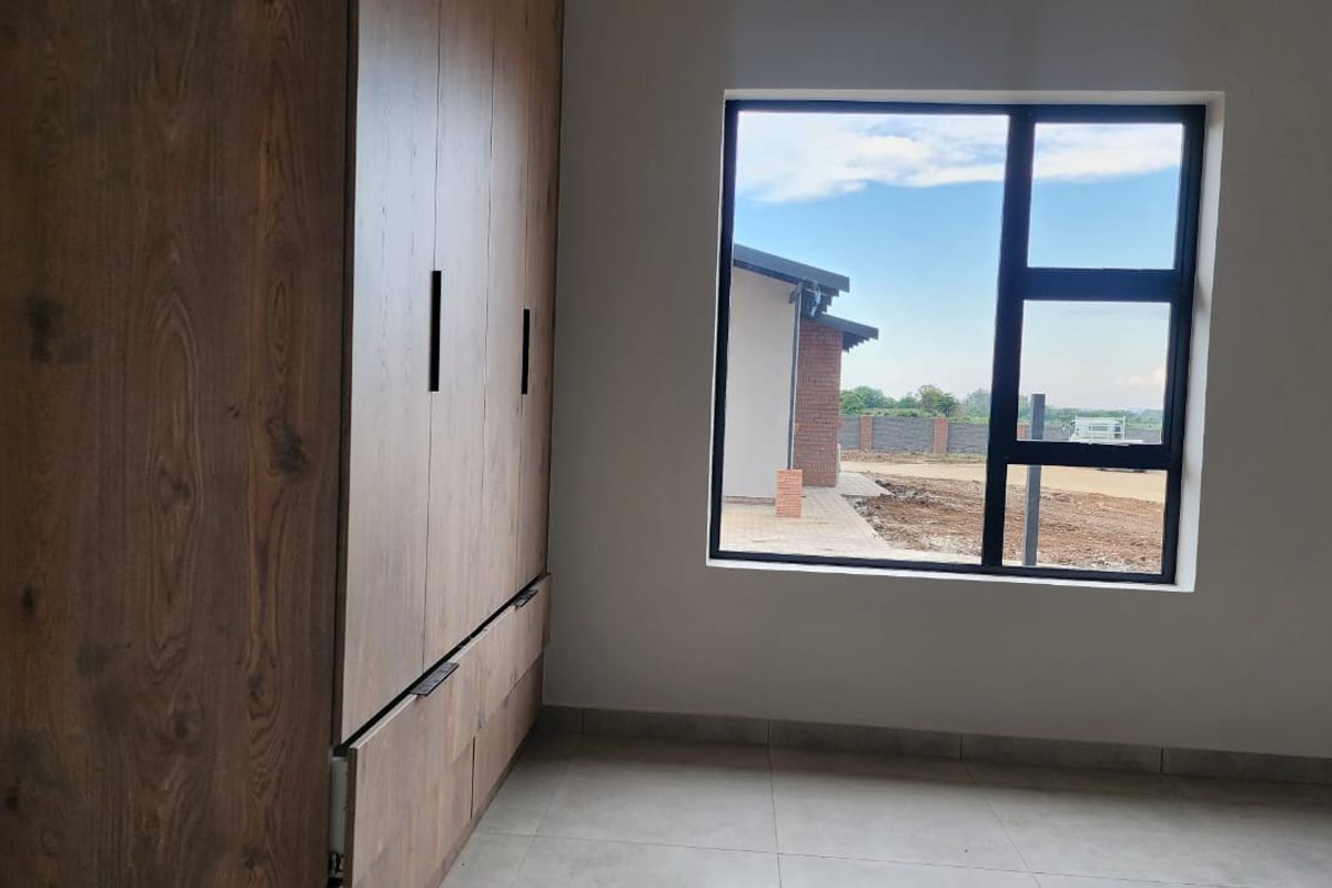 A bright, modern bedroom featuring neutral tiled floors, built-in wooden cupboards, and a large window with views of the exterior grounds.