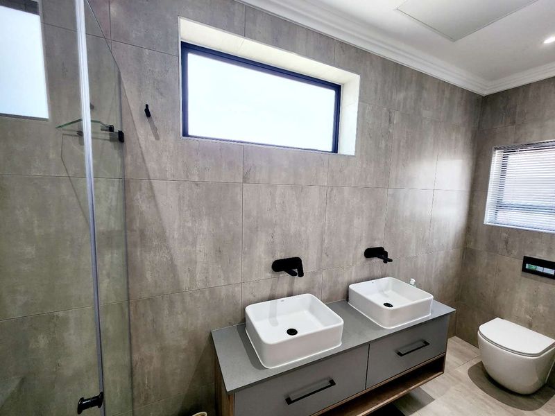 Modern bathroom featuring a double vanity with vessel sinks, wall-mounted matte black faucets, a walk-in shower, and a wall-hung toilet with grey tiling.