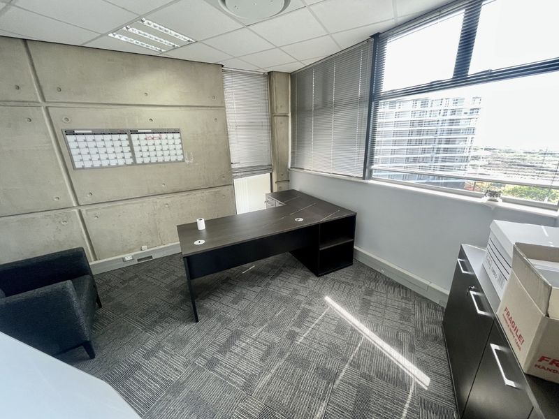 A professional office space featuring exposed concrete walls, carpeted flooring, and large windows with venetian blinds, providing ample natural light.