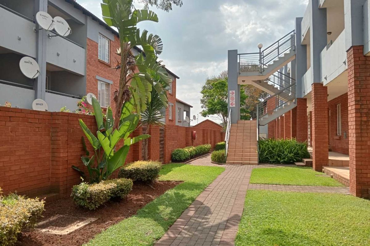 A well-maintained apartment complex courtyard featuring a paved walkway, landscaped garden beds, and external staircases providing access to upper-level units.