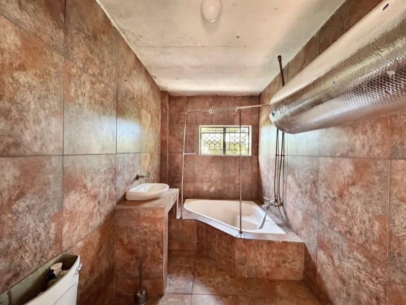 A dated bathroom featuring full-height brown ceramic wall tiles, a built-in corner bathtub, and a tiled vanity with a basin.