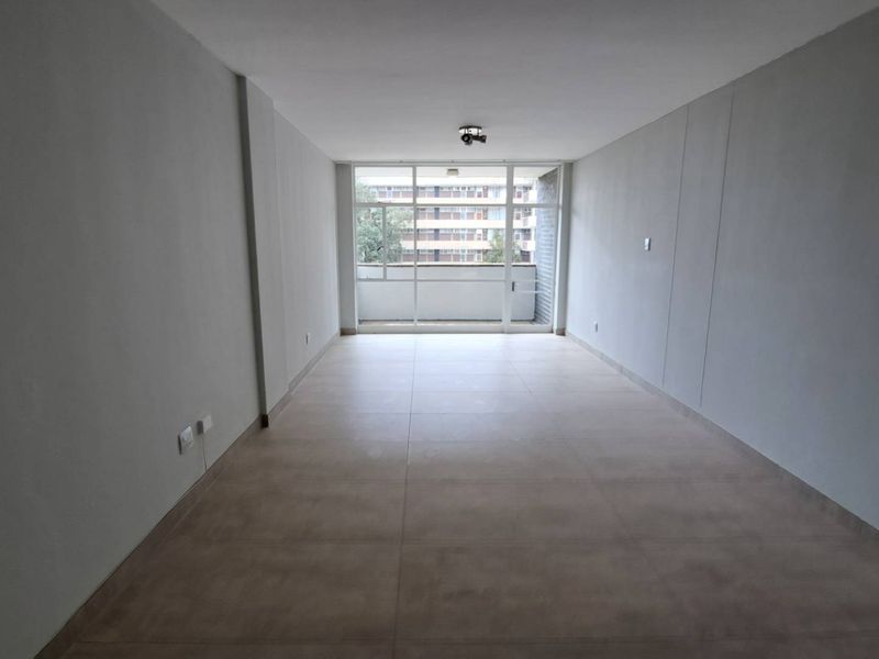 A bright, empty living room featuring neutral tiled flooring and a large sliding glass door leading to a balcony.