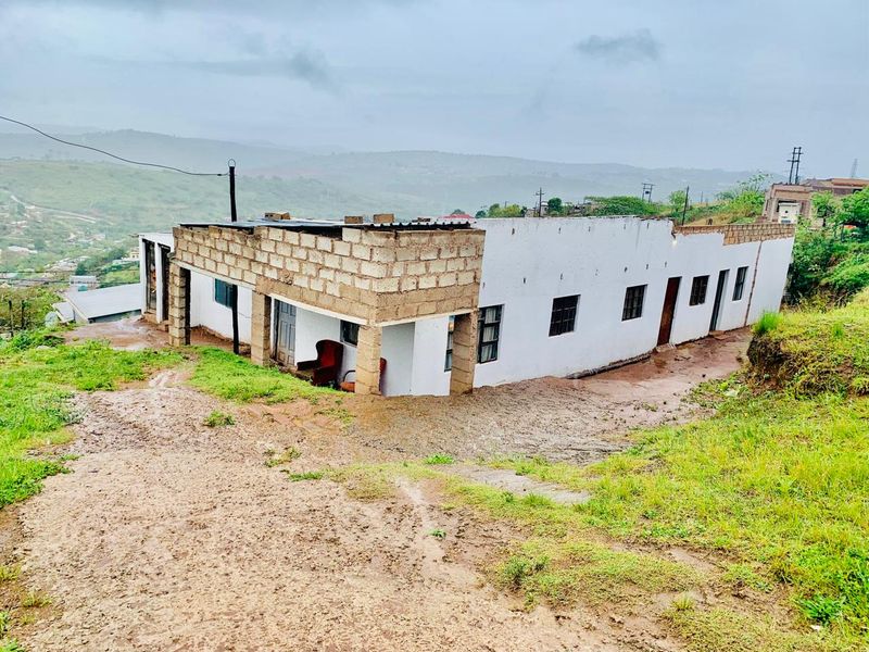 Exterior view of a single-story residential property featuring concrete block construction and a corrugated metal roof, situated on an unpaved, sloped site.