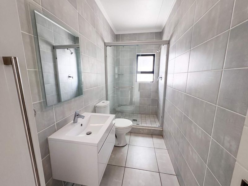 Modern bathroom featuring grey wall and floor tiles, a white vanity with basin, a toilet, and a glass-enclosed shower.