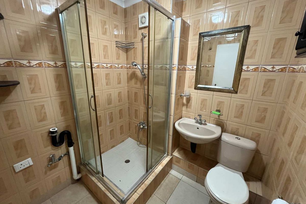 Compact bathroom featuring a shower enclosure, wall-mounted basin, and toilet, finished with dated wall tiling and exposed plumbing elements.