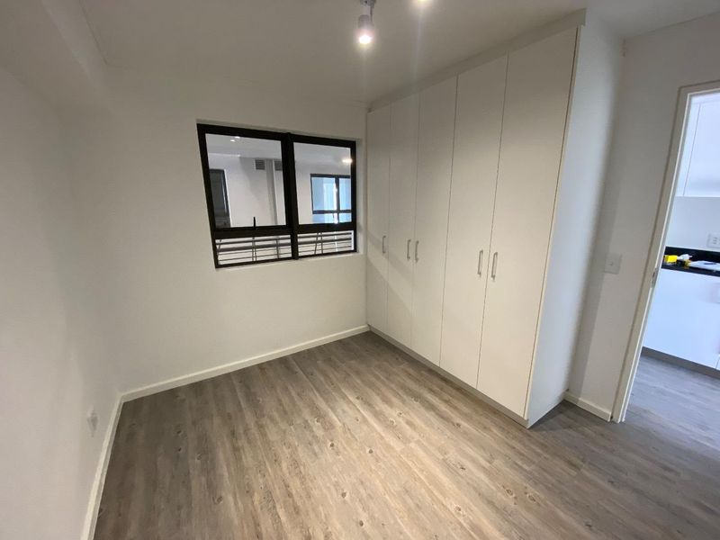 A bright, empty bedroom featuring modern laminate flooring, built-in white cupboards, and an aluminium-framed window, offering a clean and neutral living space.