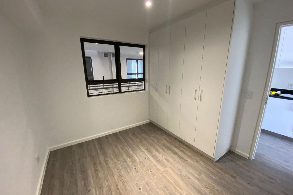 A bright, empty bedroom featuring modern laminate flooring, built-in white cupboards, and an aluminium-framed window, offering a clean and neutral living space.