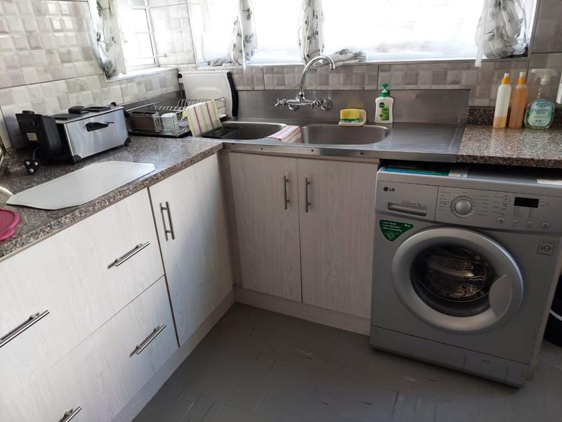 A functional laundry area featuring light wood-grain cabinetry, a stainless steel double sink, granite-look countertops, and space for a front-loading washing machine.