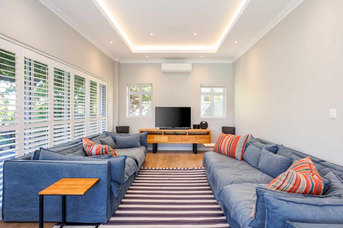 A bright and modern living room featuring timber flooring, recessed cove lighting, split-system air conditioning, and white plantation shutters.
