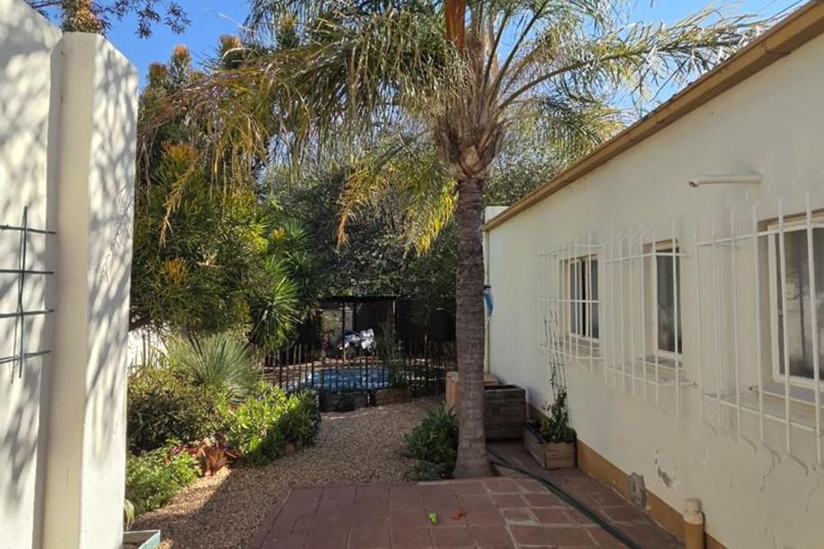 Rear exterior view featuring a tiled patio, security bars on windows, gravel garden area, and a swimming pool in the background.