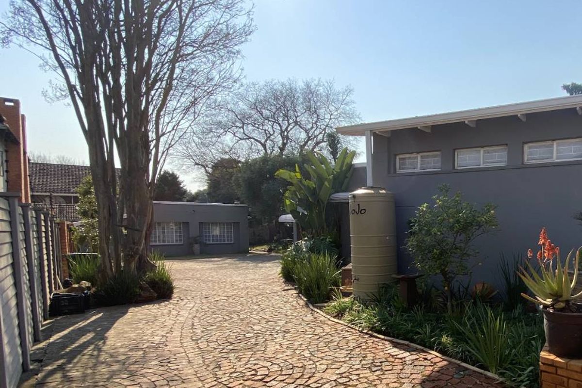 A well-maintained rear exterior area featuring a cobblestone driveway, established garden beds, and a water tank alongside the building structure.