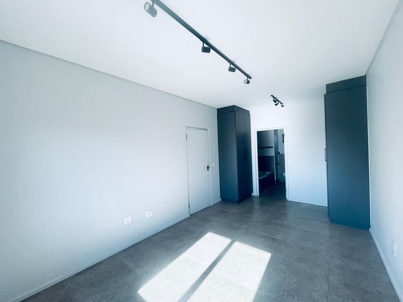 A modern, empty bedroom featuring sleek dark built-in cupboards, contemporary track lighting, and neutral tiled flooring, with an adjoining bathroom visible.