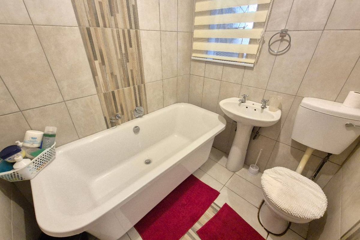 A functional bathroom featuring a freestanding bathtub, pedestal basin, and toilet, finished with neutral wall tiles and a decorative mosaic accent strip.
