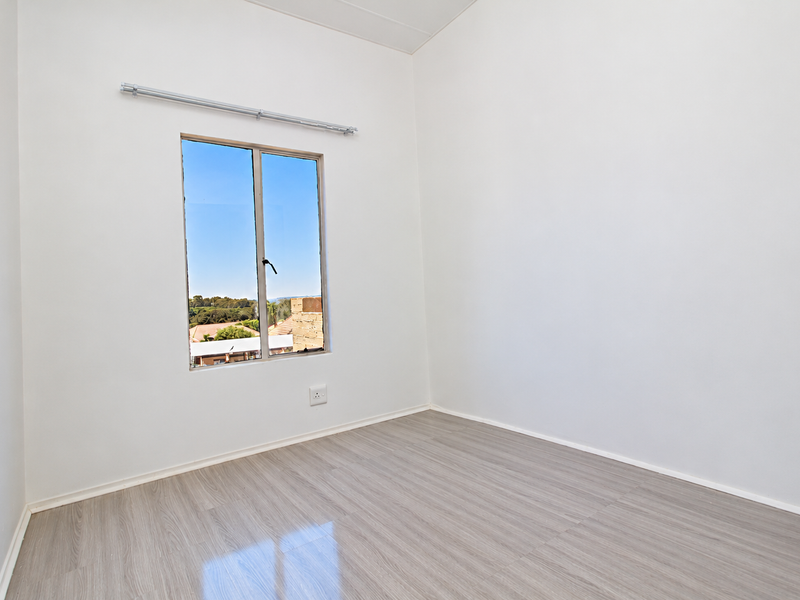 A bright, empty bedroom featuring light-toned laminate flooring, a single window with a curtain rod, and neutral white walls.