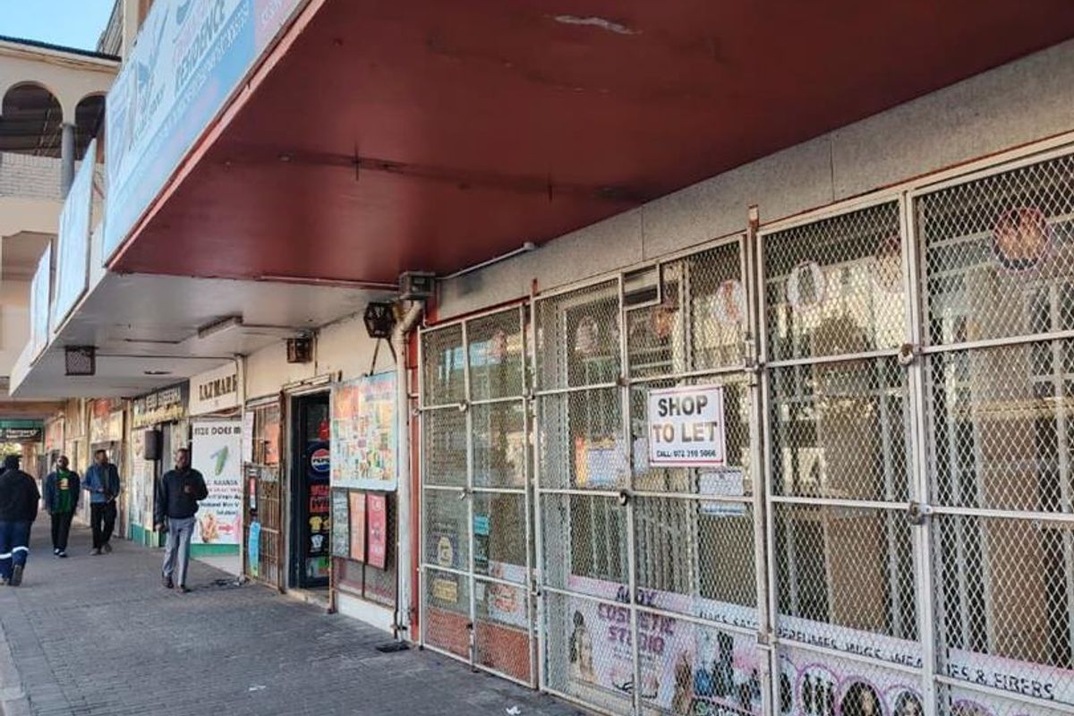 Street-level view of a commercial shopfront featuring metal security gates, signage, and a covered walkway in a busy urban retail area.