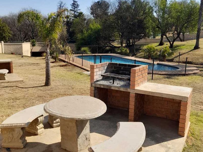 Outdoor communal pool area featuring a built-in brick braai, concrete seating, and a fenced swimming pool set within a landscaped garden.