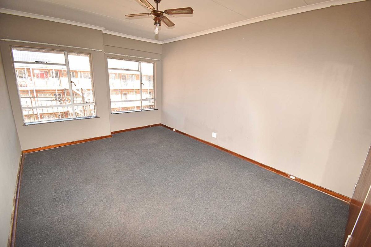 An empty bedroom featuring dark carpeting, large windows providing natural light, and a ceiling fan, offering a blank canvas for renovation.