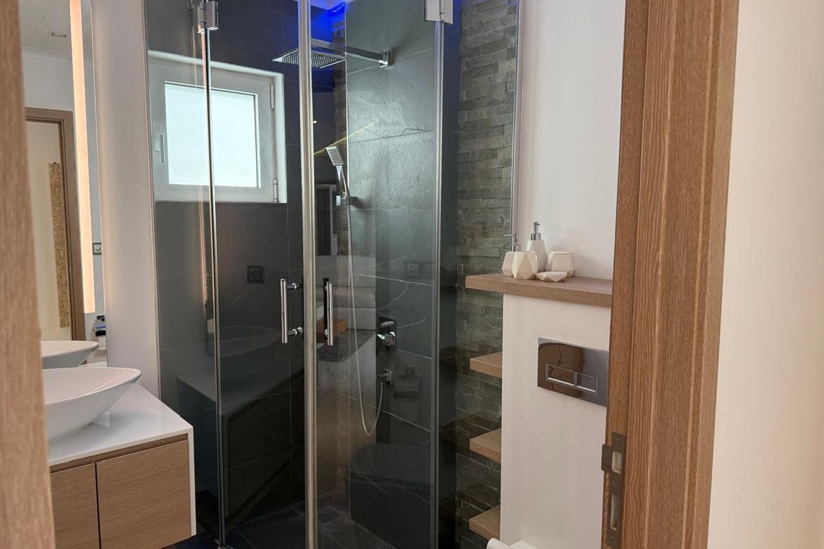 Modern bathroom featuring a glass-enclosed walk-in shower with stone-tiled accent wall, floating vanity with vessel sink, and wall-mounted toilet flush plate.