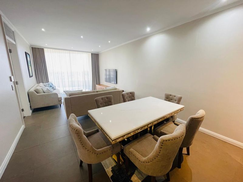 A spacious open-plan living and dining area featuring neutral tiled flooring, recessed downlighting, and large windows with floor-to-ceiling curtains.