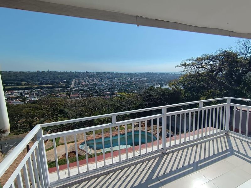 A spacious elevated balcony featuring a white metal balustrade and tiled flooring, offering expansive views over the neighborhood and swimming pool area.