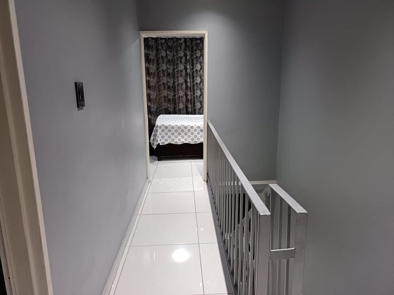 A bright upper-level hallway featuring white tiled flooring and a metal balustrade overlooking the staircase, leading into an adjacent bedroom.