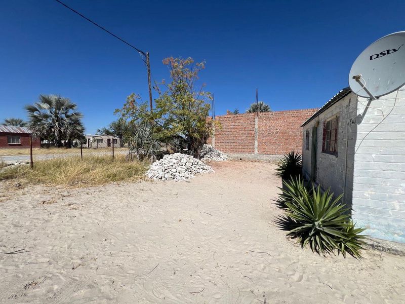 Rear exterior view showing a sandy yard, unfinished brick boundary wall, and existing residential structure with a satellite dish.