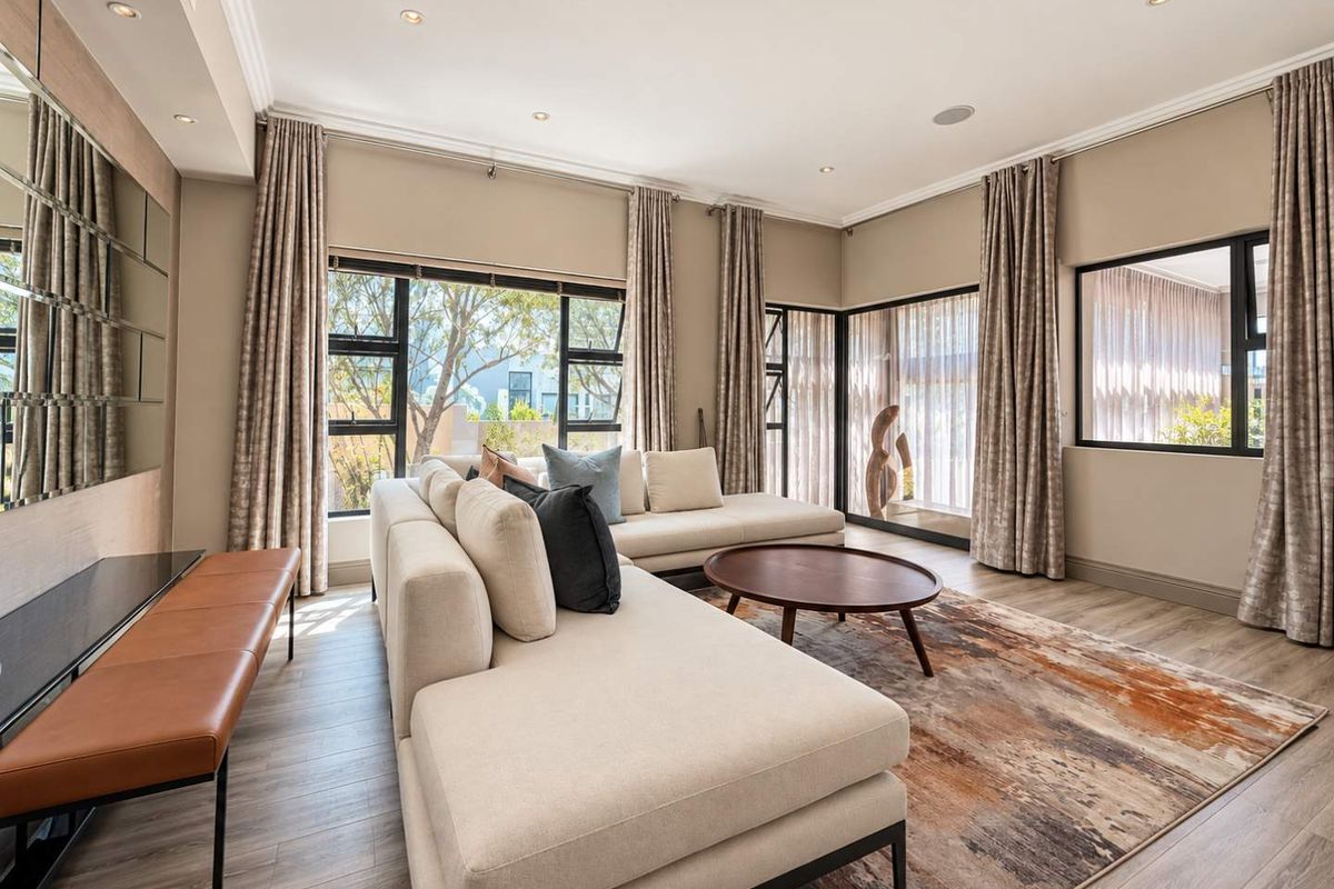 A bright and modern living room featuring large aluminium-framed windows, recessed ceiling lighting, integrated speakers, and elegant mirrored wall accents.