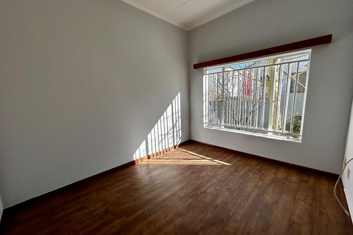 A bright, empty bedroom featuring laminate flooring, a large window with security bars, and a neutral wall finish.
