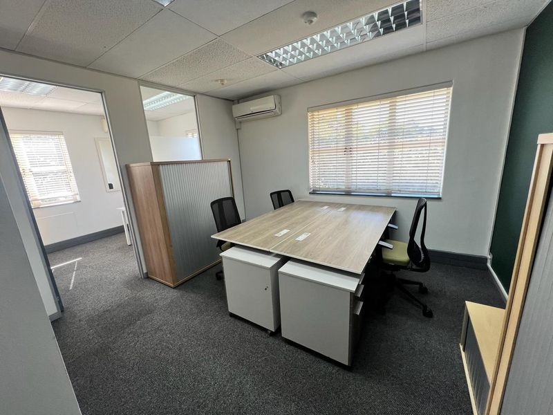 A functional office space featuring carpeted flooring, a suspended ceiling with recessed lighting, split-system air conditioning, and large windows with venetian blinds.