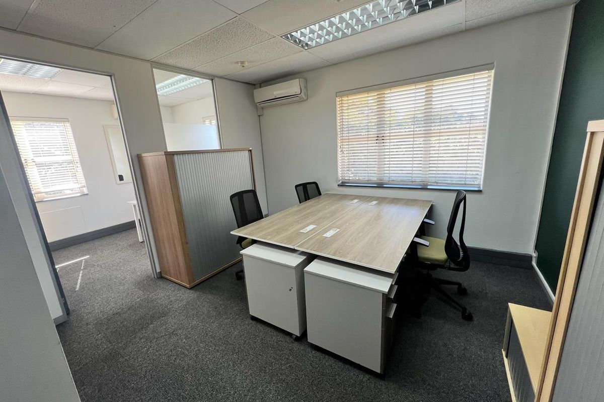 A functional office space featuring carpeted flooring, a suspended ceiling with recessed lighting, split-system air conditioning, and large windows with venetian blinds.
