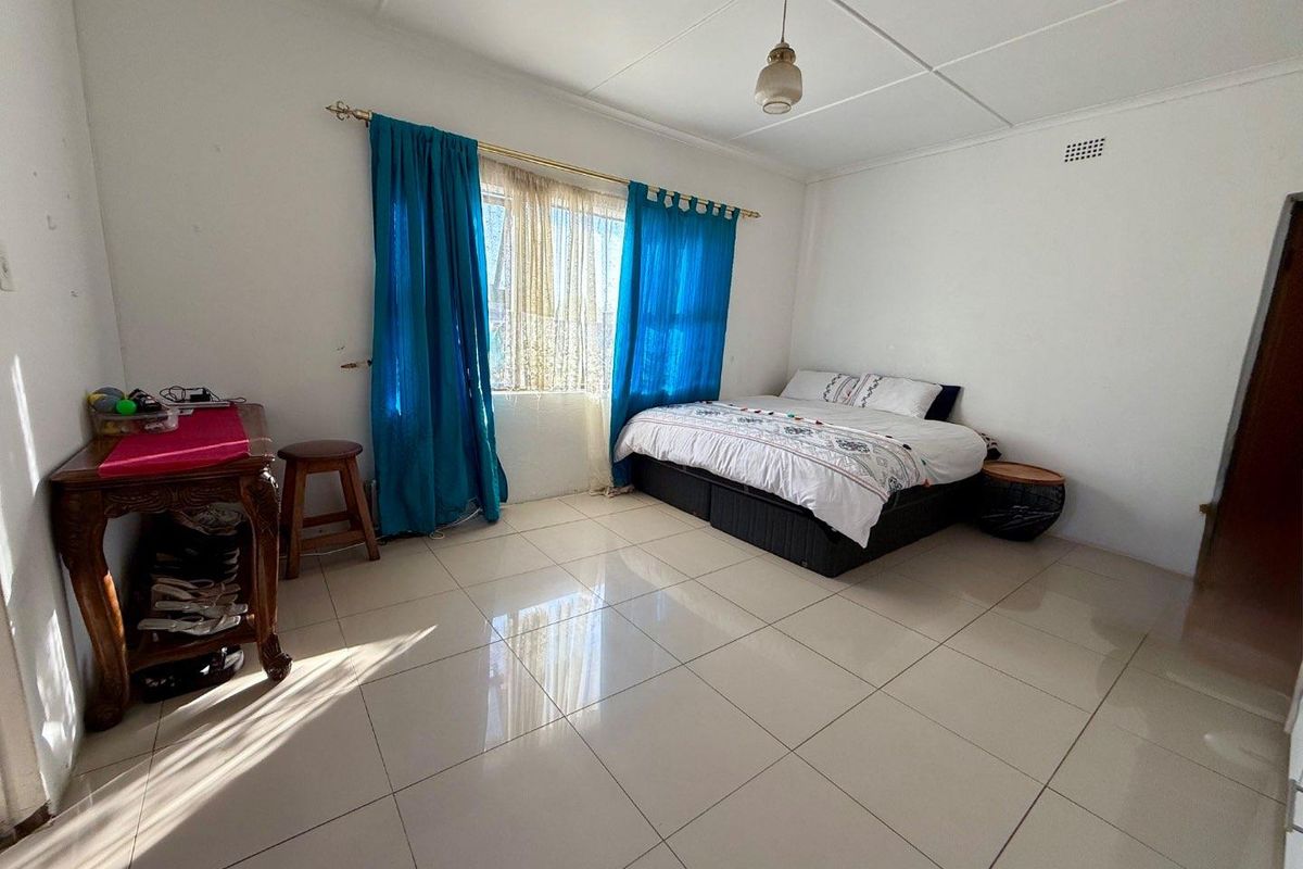 A spacious bedroom featuring large neutral floor tiles, a simple ceiling design, and a window with curtain fixtures.