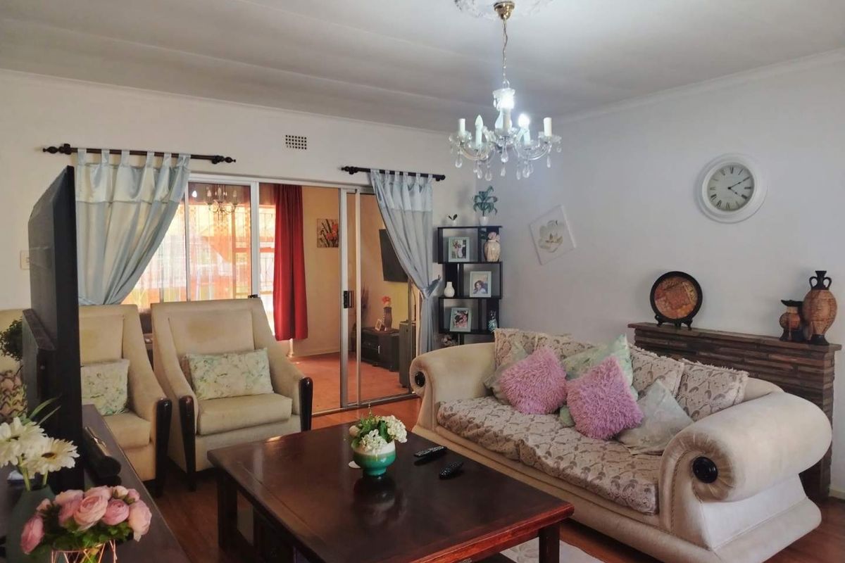 A comfortable living room featuring a classic chandelier, ceiling rose, and sliding glass doors leading to an adjacent space, with warm laminate flooring throughout.