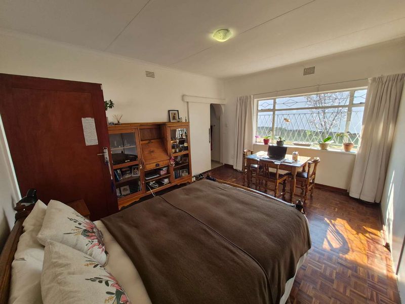 A spacious bedroom featuring classic parquet flooring, large windows with security bars, and neutral wall finishes, offering a bright and functional living space.