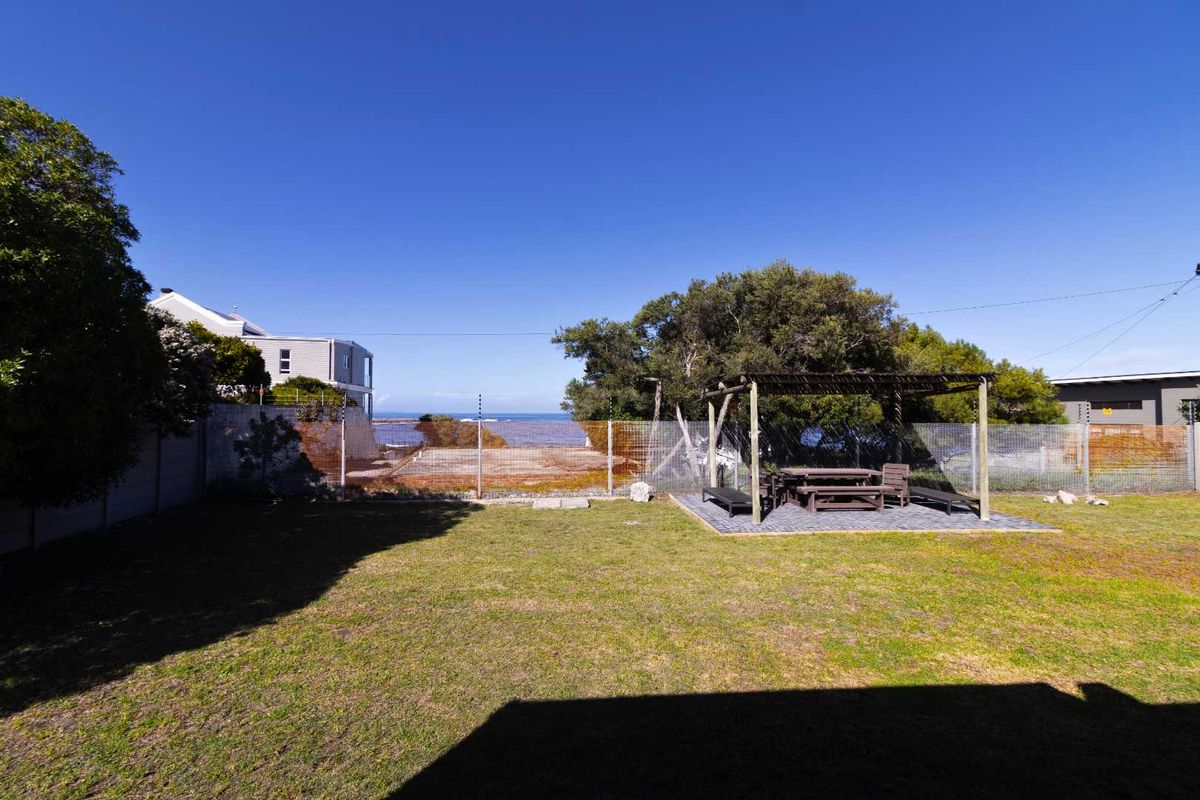 A spacious rear garden featuring a paved patio area with a wooden pergola, enclosed by wire fencing with distant ocean views.