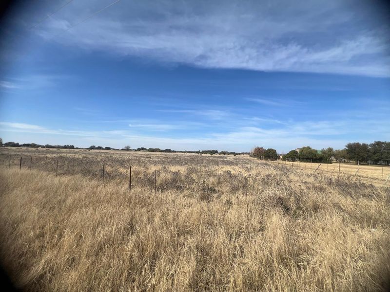 A wide, open residential vacant land plot featuring dry grass and perimeter wire fencing under a clear blue sky.