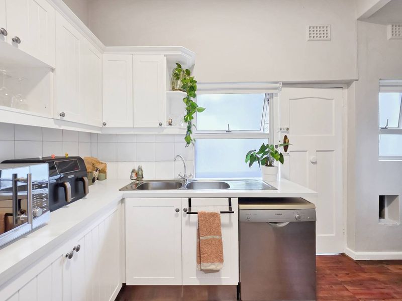 A bright, functional kitchen featuring white cabinetry, a double stainless steel sink, tiled splashback, and timber flooring, offering a clean and well-maintained workspace.