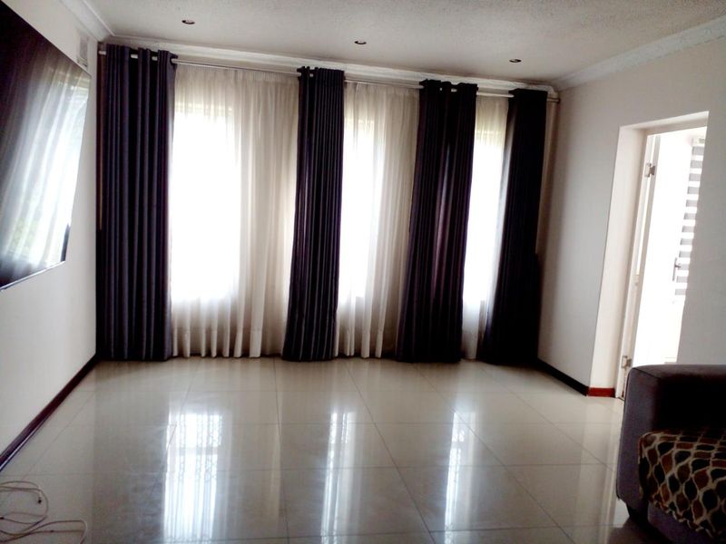 A spacious, empty living room featuring polished tiled flooring, recessed downlighting, and large windows dressed with floor-to-ceiling curtains.